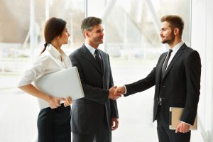 Two businessmen shaking hands next to a businesswoman carrying a white laptop