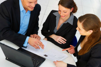 Three business co-workers around a laptop viewing some paper documents