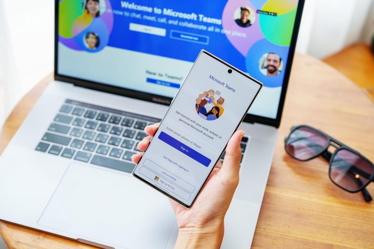 Person holding a phone showing the Microsoft Teams sign-in screen in front of a Teams laptop