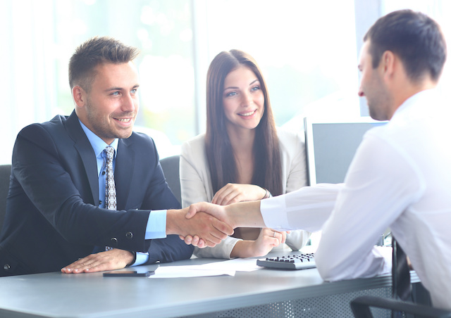 Two businessmen shaking hands while a woman smiles in the background