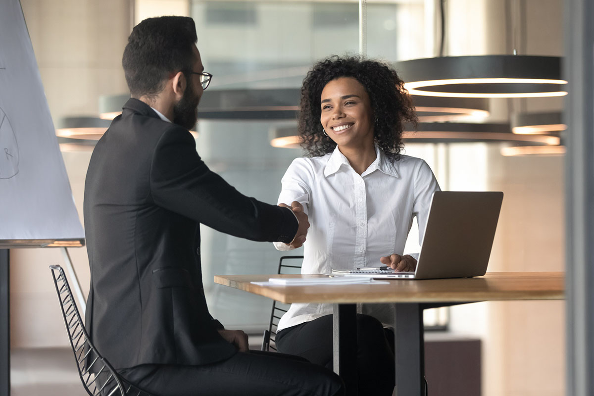 Two professionally dressed people sitting and shaking hands over a table with a laptop on it