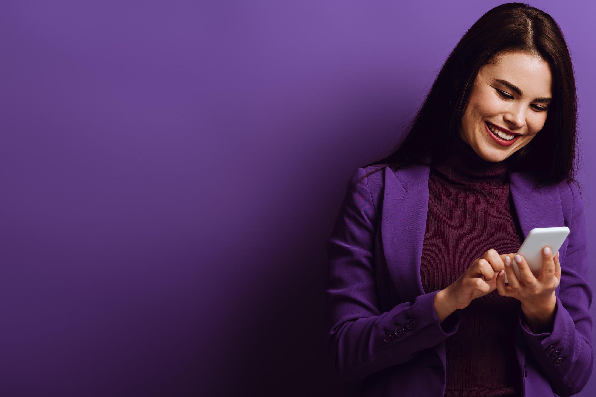 Cheerful young woman smiling while chatting on smartphone, against purple background